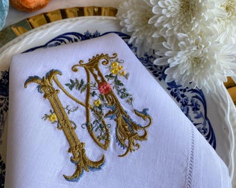 May include: White linen napkin with an embroidered gold letter "M" adorned with floral accents in yellow, pink, and blue. The napkin is placed on a blue and white patterned plate, with decorative pumpkins and white flowers in the background.