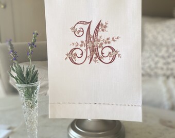 May include: White linen hand towel with an embroidered burgundy letter "M" and floral accents. The towel is hanging on a silver towel rack. A bar of soap with a floral design and a vase of lavender are in the foreground.