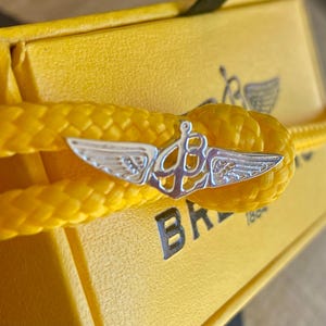 May include: A yellow box with a yellow rope bracelet with a silver winged logo. The logo says "BREITLING 1884".