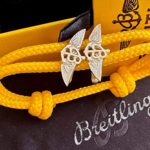 May include: A yellow braided rope bracelet with silver-colored metal airplane wing charms. The bracelet is knotted and rests on a black surface with the word "Breitling" in silver script. A yellow box is in the background.