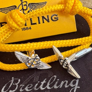 Op de afbeelding: Een gele touw armband met zilverkleurige vliegtuigvormige manchetknopen. De armband is bovenaan geknoopt. Een gele doos met het woord "Breitling" en het jaartal "1884" staat op de achtergrond. De manchetknopen liggen op een zwarte ondergrond.