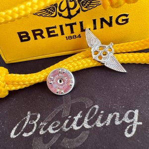 May include: A silver-colored Breitling pin with wings, a yellow rope, and a yellow box with the Breitling logo. The pin is next to a silver-colored round object with text. The box has the text "Breitling 1884".