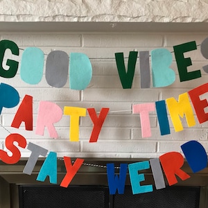 May include: A felt banner with the words "Good Vibes Party Time Stay Weird" in various colors. The banner is strung up on a white brick wall above a fireplace.