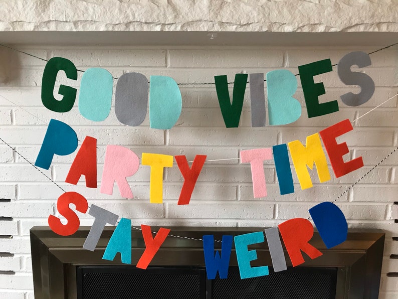 May include: A felt banner with the words "Good Vibes Party Time Stay Weird" in various colors. The banner is strung up on a white brick wall above a fireplace.