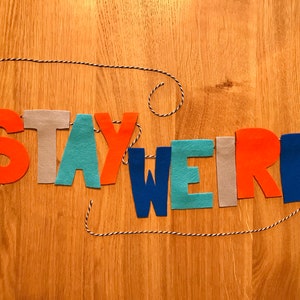 May include: A felt banner with the words "STAY WEIRD" in colorful letters. The letters are strung on a white and black twine.