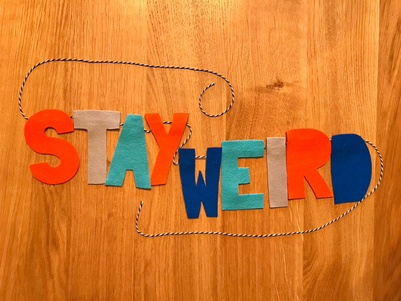 May include: A felt banner with the words "STAY WEIRD" in colorful letters. The letters are strung on a white and black twine.