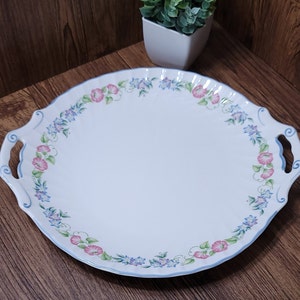 Puede incluir: Plato de servir de cerámica blanca con borde festoneado y dos asas. El plato está decorado con un patrón floral azul y rosa.
