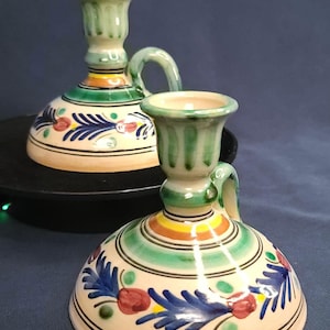 May include: Two ceramic candle holders with a cream base, green, blue, red, and yellow accents. Each has a handle and is decorated with floral patterns. The candle holders are on a black surface against a dark blue background.