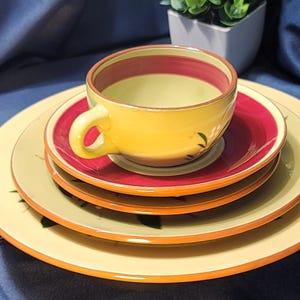 Puede incluir: Una taza de té amarilla con interior rojo y amarillo se encuentra sobre una pila de platos a juego. Los platos y la taza tienen un diseño floral y un borde naranja. La taza tiene un asa curva. El juego está sobre una superficie azul.