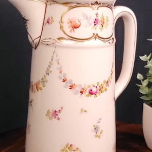 May include: A white porcelain jug with a lid, decorated with hand-painted floral patterns in pink, yellow, and blue. Gold trim highlights the rim, handle, and floral details. The jug has a tall, slender form.