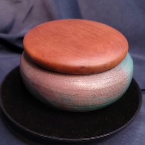 May include: A handcrafted wooden box with a round, reddish-brown lid. The body of the box is a muted pink and teal color, sitting on a black, circular base. The box is likely used for storage.