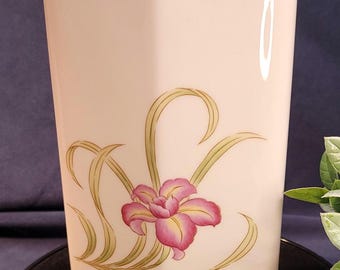 May include: A cream-colored, hexagonal vase with a gold rim and a floral design. The design features a pink and yellow flower with green leaves. The vase is on a black surface against a dark blue background.