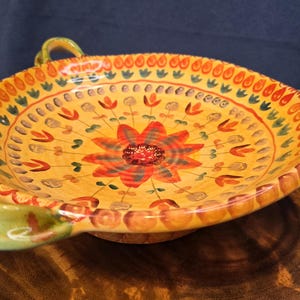 May include: A round ceramic bowl with two handles, featuring a vibrant yellow base color decorated with a central red flower motif, surrounded by concentric rings of small painted floral patterns and orange teardrop shapes along the rim.