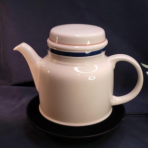 Puede incluir: Una tetera de cerámica blanca con tapa, boquilla y asa. La tapa tiene un diseño de rayas rosas y azules. La tetera está sobre un platillo negro. El fondo es azul oscuro.