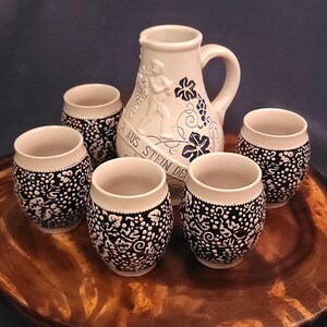 May include: A cream-colored ceramic pitcher and five matching small cups. The pitcher has a handle and a relief design of a figure and grapes. The cups and pitcher are decorated with a black and white grape and vine pattern.