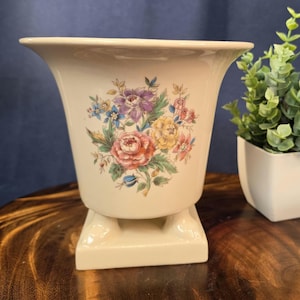 May include: A cream-colored ceramic vase with a flared rim and a square pedestal base. The front of the vase features a colorful floral decal with pink, yellow, purple, and blue flowers surrounded by green leaves on a smooth, glossy surface.