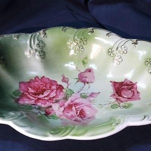 May include: A white porcelain serving bowl with a green and white floral design. The bowl has two handles and is decorated with pink roses and gold accents.