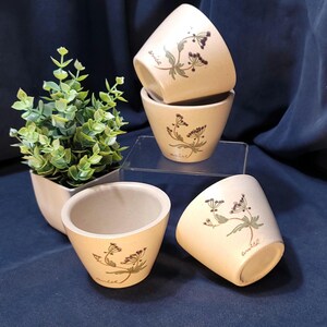 May include: Set of four small, beige ceramic cups with a floral design and the word "anise" written on each cup.