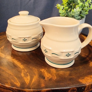 May include: A cream-colored ceramic sugar bowl with a lid and a matching creamer pitcher. Both pieces feature a raised basket-weave texture around the center, accented with small green floral motifs and a thin green band near the base.