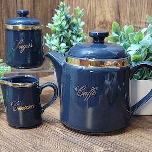 May include: Three ceramic kitchen canisters in a dark blue color with gold trim. The canisters are labeled "Sugar", "Cream", and "Caffe".