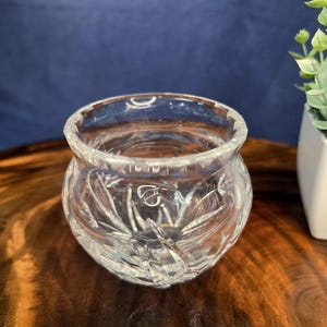 May include: A clear, round crystal bowl with a detailed, etched pattern. The bowl has a wide opening and sits on a wooden surface. The background is a solid blue color. The bowl is likely used for decorative purposes.