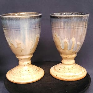 May include: Two handmade ceramic goblets. The base and stem are speckled light beige, with the upper portion transitioning from light beige to light blue and a dark blue rim. The goblets are displayed on a dark surface.
