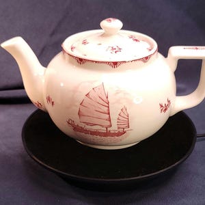 May include: White ceramic teapot with a red sailboat design and floral accents. The teapot has a matching lid and a square handle. It sits on a black, round base.