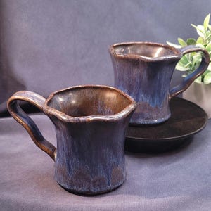 May include: Two handcrafted ceramic mugs with a distinctive, wavy rim. The mugs feature a deep blue glaze with brown highlights, giving them a rustic, artistic look. The mugs are on a black plate, ideal for tea or coffee.