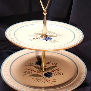 Puede incluir: Bandeja de dos niveles con asa y soporte dorados. Los platos blanquecinos presentan un borde festoneado azul y un diseño floral con detalles azules y rojos. Una pieza decorativa para servir postres o aperitivos.