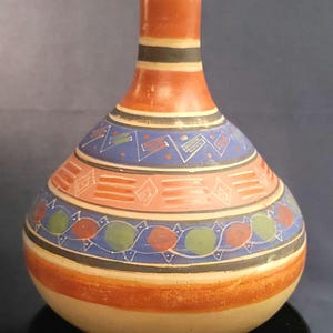 May include: A hand-painted ceramic vase with a rounded body and a long neck. The vase features a cream-colored base, an orange neck, and bands of blue, red, and green geometric patterns. The vase is approximately 8 inches tall.