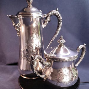 Op de afbeelding: Een zilveren theeservies met sierlijke bloemengravures. De set bevat een hoge theepot en een kleinere room- of suikerpot, beide met decoratieve handvatten en deksels. De set staat op een zwarte schaal tegen een donkerblauwe achtergrond.