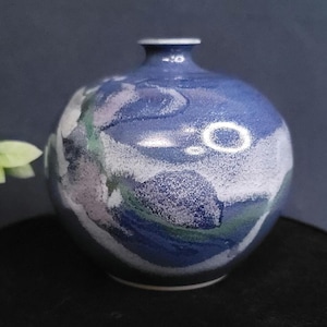 May include: A round, blue ceramic vase with a narrow neck. The vase features a speckled design with white, green, and purple accents. The vase has a glossy finish and sits on a dark surface.