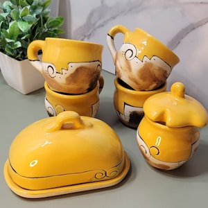 Könnte beinhalten: Ein Set gelber Keramik-Küchenutensilien mit braunen und weißen Akzenten. Das Set enthält vier Tassen, eine Butterdose und ein kleines Glas mit Deckel.