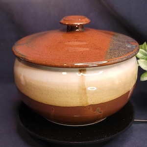 Puede incluir: Una olla de cerámica con tapa de forma redonda. La tapa y la banda inferior son de color marrón rojizo, mientras que la sección media es crema y beige. La olla se asienta sobre una base redonda negra.