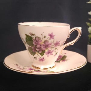 May include: A vintage teacup and saucer set. The cup and saucer are white with a floral design of purple violets and green leaves. Gold trim accents the edges of the cup and saucer.