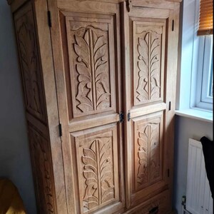 May include: A tall, wooden wardrobe with two doors and a drawer. The doors are carved with intricate leaf designs. The wardrobe is made of light brown wood and has a distressed finish.