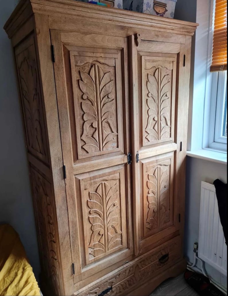 May include: A tall, wooden wardrobe with two doors and a drawer. The doors are carved with intricate leaf designs. The wardrobe is made of light brown wood and has a distressed finish.