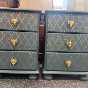 May include: Two blue bedside tables with three drawers each. The drawers have gold elephant-shaped knobs and a gold geometric pattern.