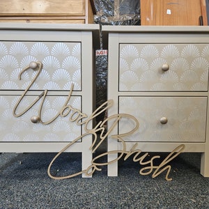 May include: Two beige bedside tables with two drawers each. The drawers feature a white geometric pattern. The bedside tables are made of wood and have gold-coloured hardware. The words "Wood & Brush Co." are written in gold on the floor in front of the bedside tables.