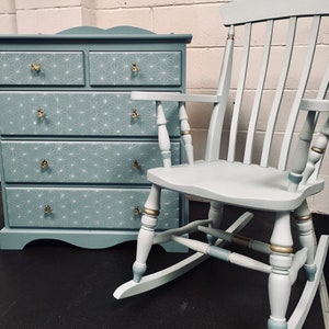 May include: A light blue chest of drawers with five drawers and gold knobs, paired with a light blue rocking chair with gold accents. The dresser has a geometric pattern on the front.