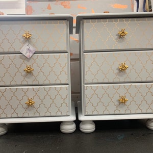 May include: Two white bedside tables with three drawers each. The drawers have gold bee-shaped knobs and a gold geometric pattern.