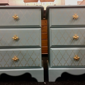 May include: Two light blue bedside tables with three drawers each. The drawers have gold bee-shaped knobs and a gold geometric pattern.