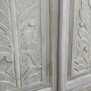 May include: Two white painted wooden doors with carved leaf designs. The doors are closed and the edges of the doors are visible.
