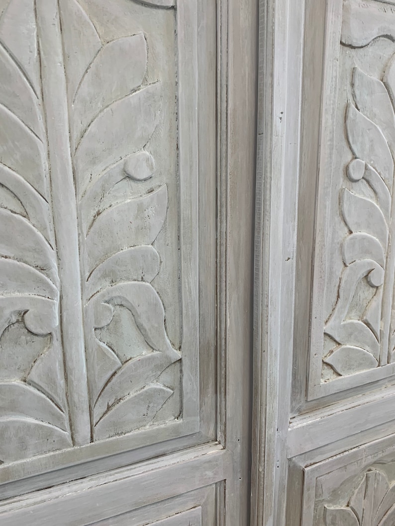 May include: Two white painted wooden doors with carved leaf designs. The doors are closed and the edges of the doors are visible.
