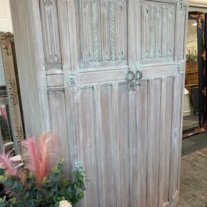 May include: A whitewashed wooden armoire with intricate carvings and ornate hardware. The armoire has two doors and is standing on a concrete floor.