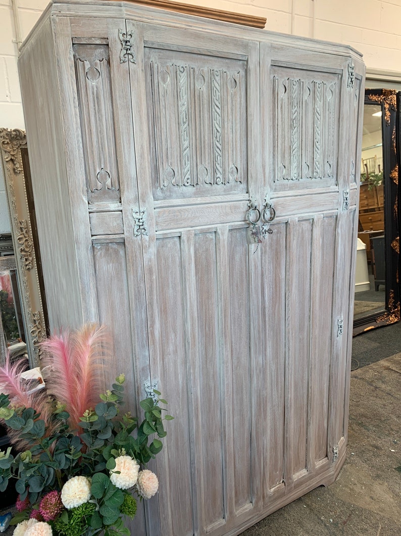 May include: A whitewashed wooden armoire with intricate carvings and ornate hardware. The armoire has two doors and is standing on a concrete floor.