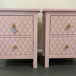 May include: Two matching pink bedside tables with two drawers each. The drawers have a gold geometric pattern and gold bee-shaped knobs.