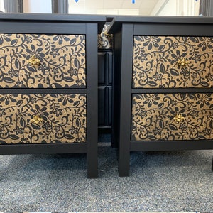 May include: Two black bedside tables, each with two drawers. The drawer fronts display a gold and black floral design. Each drawer has a gold-coloured knob. The tables are set against a neutral background.