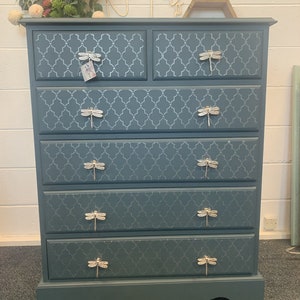 May include: A teal blue chest of drawers with six drawers. Each drawer has a silver dragonfly pull and a white geometric pattern.