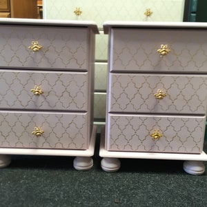May include: Two matching pink bedside tables with three drawers each. The drawers have gold bee-shaped knobs and a gold geometric pattern.